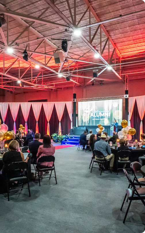 Individuals seated at tables watching stage with “ALUMNI” sign in decorated event hall; drapes and gold balloons present.