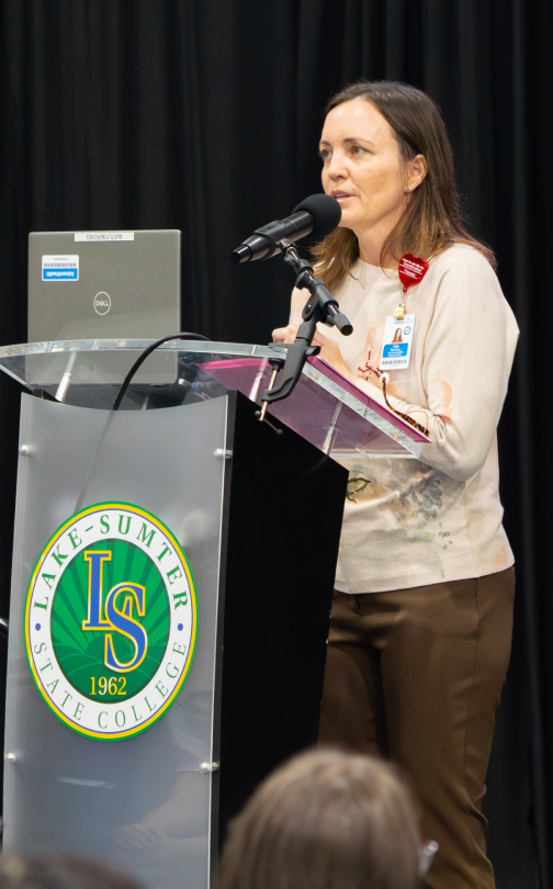 Instructor at podium with Lake-Sumter State College emblem, laptop, and microphone; likely classroom or lecture hall setting.