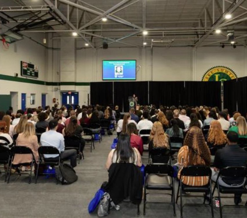 Individuals seated in rows facing person at podium in gymnasium; screen and banners visible.
