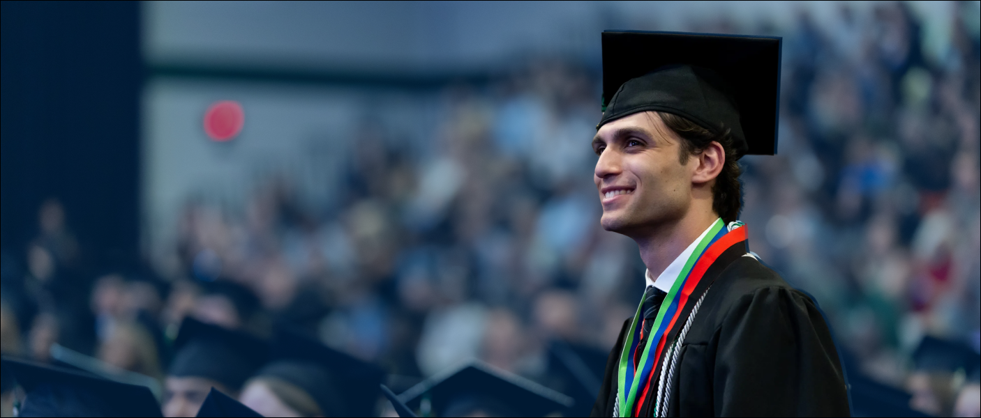 Student in cap, gown, and honor cords stands in crowded auditorium. Graduation attire present.