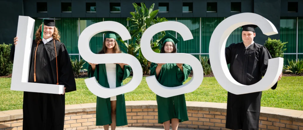 Four students in graduation attire hold large LSSC letters outdoors near a building and greenery.