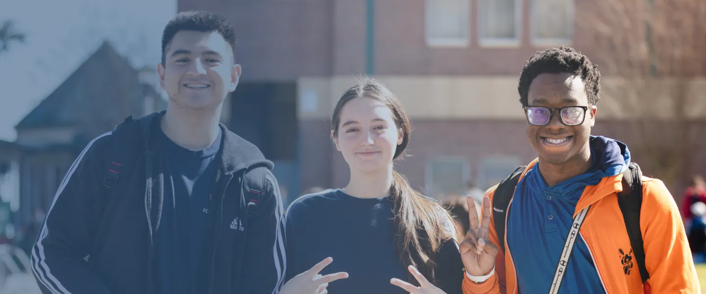 Three students outdoors, making peace signs, brick building blurred in background.
