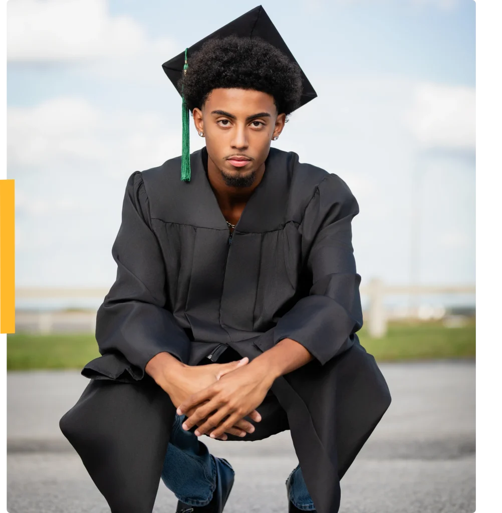 Student in black graduation gown and cap squats outdoors, looking at camera with confident expression.