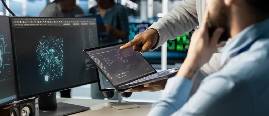 Two individuals review code and a digital brain image on computer monitor and laptop in office setting.