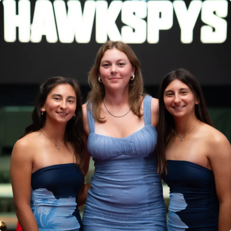 Three individuals in blue dresses pose, smiling, in front of a large HAWKSPYS sign at an indoor event.
