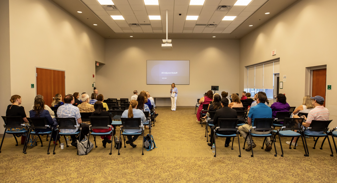 Person speaks at front of conference room to seated audience, projection screen visible.