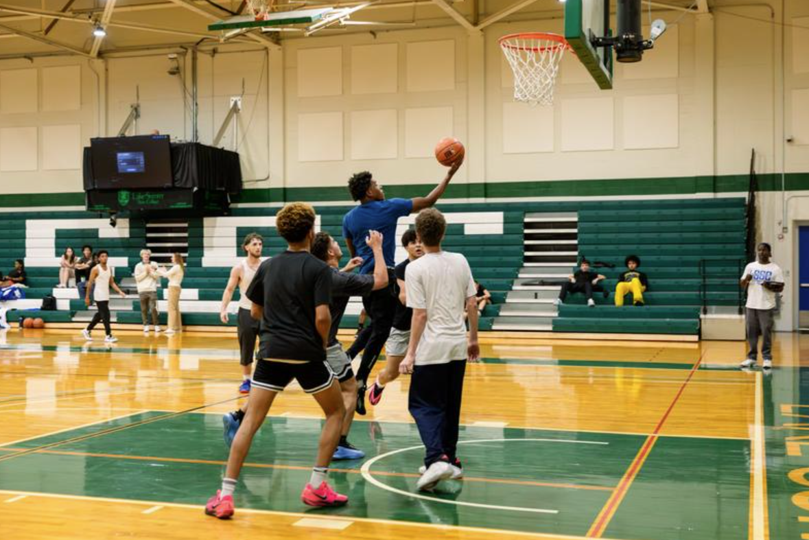 Five individuals play basketball in a gym; one person jumps, shooting the ball toward the hoop.
