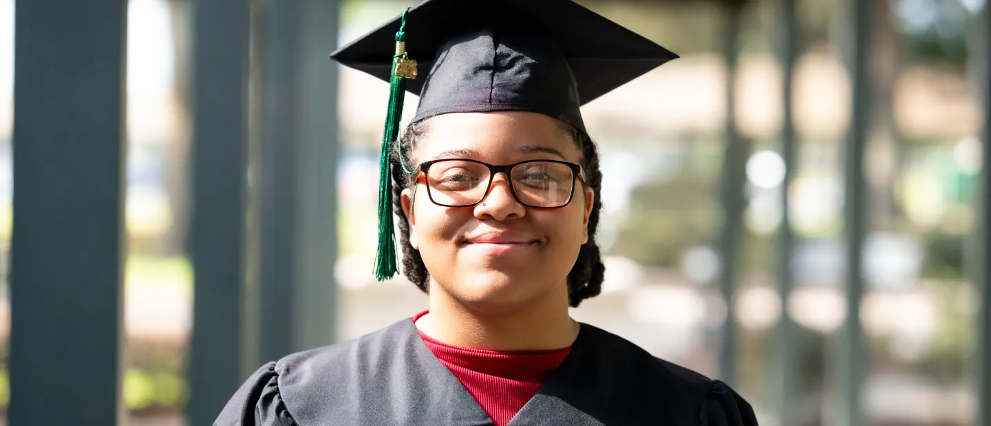 Student grad in glasses and graduation attire smiling outdoors.