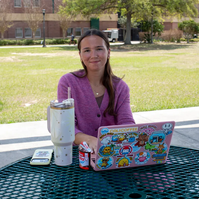 Student at outdoor table with sticker-covered laptop, drink cup, and snacks.