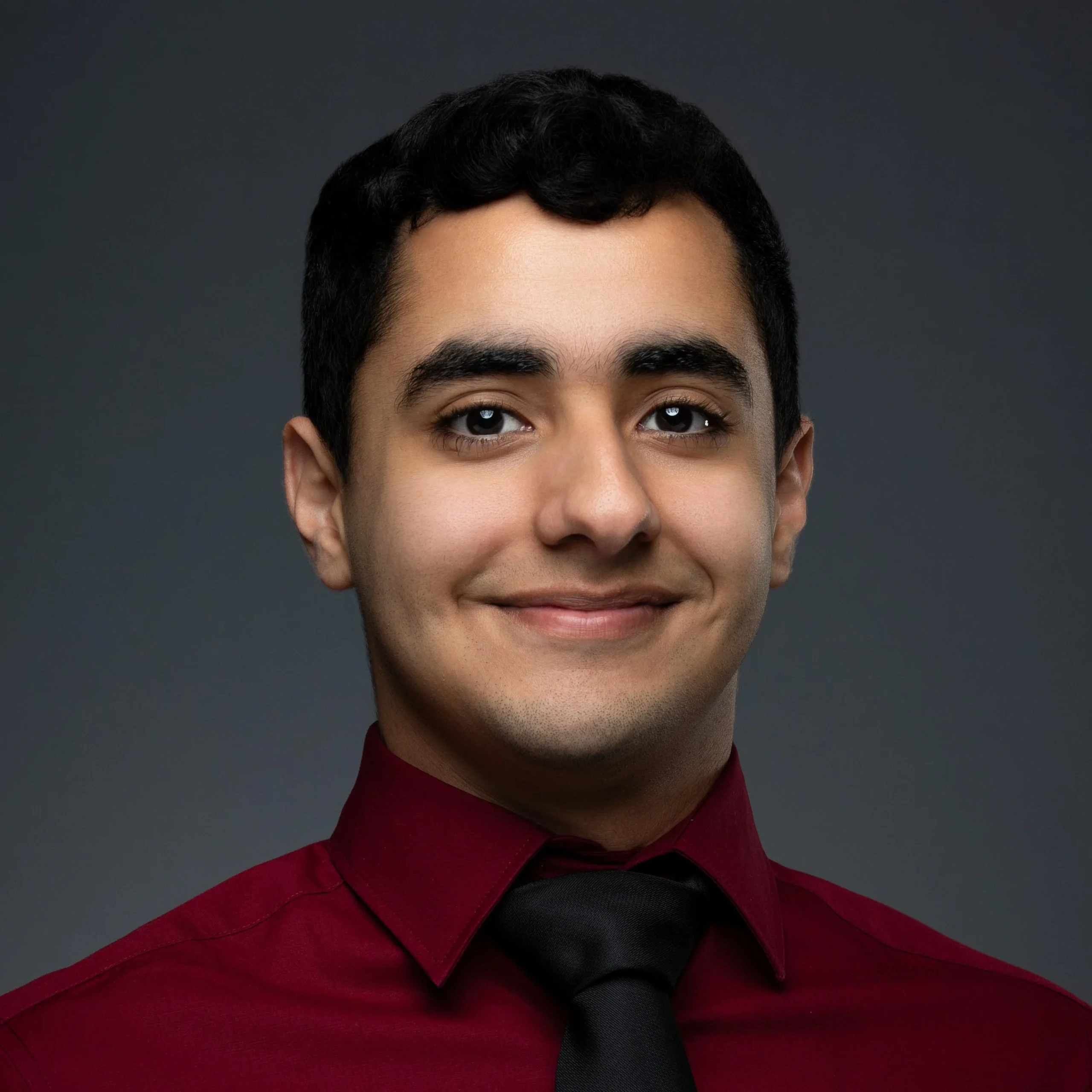 Individual in burgundy shirt and black tie smiling, plain dark background. No clear role or setting indicated.