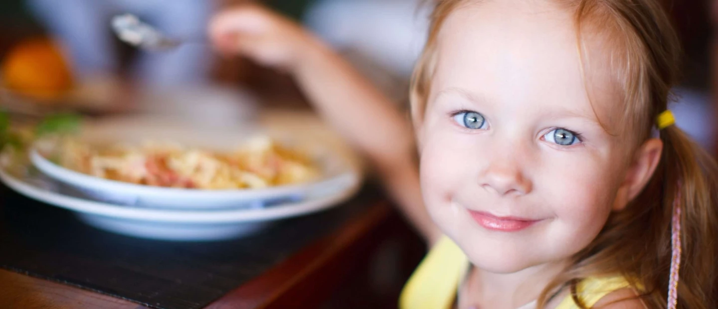 Close-up of a child eating pasta at a table. The child holds a fork with food, and a plate of pasta is in front of them