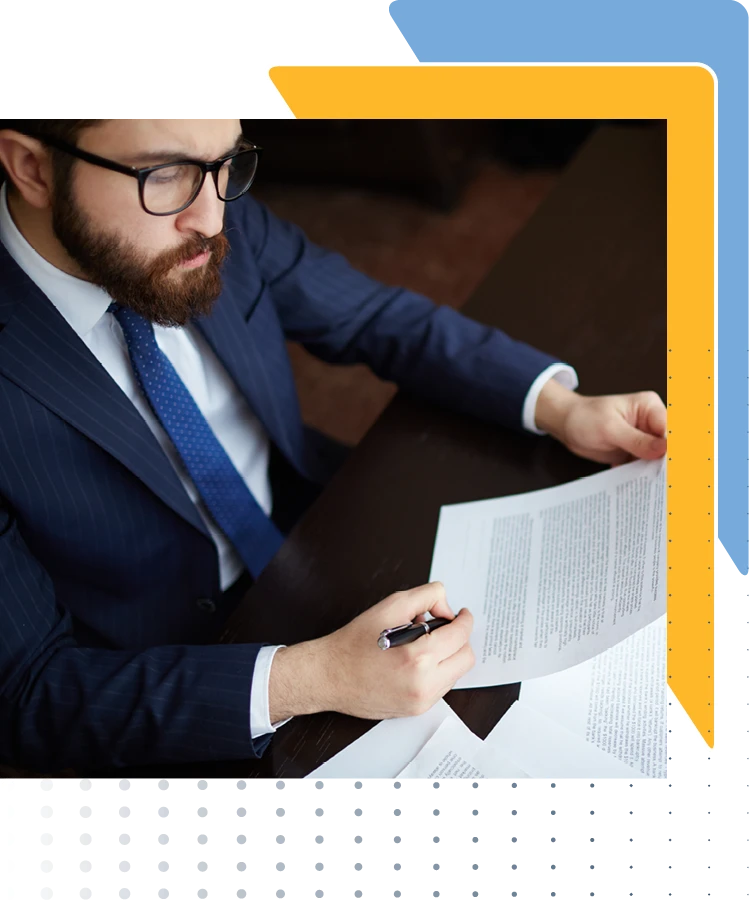 a man in a blue pinstripe suit sitting at a desk, reviewing documents. The person's face is blurred, and they are holding a pen while looking at one of the documents