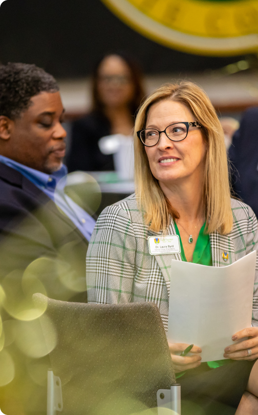 Individual in glasses holding papers, smiling next to another person at professional or academic event (setting unclear).