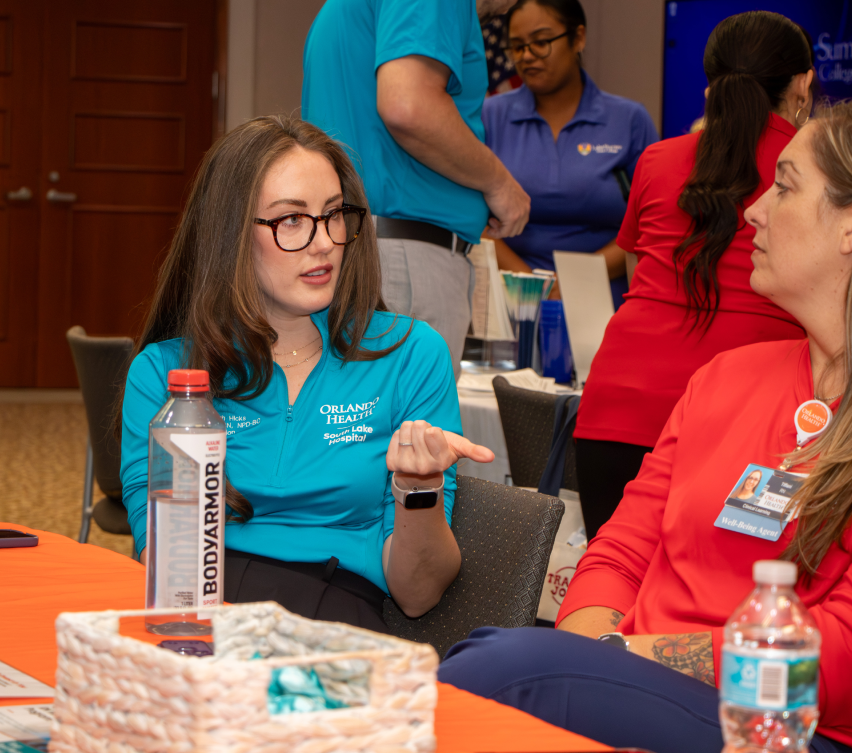 Two individuals seated at a table in the Magnolia room, conversing; one gestures. Bottled water on table. Event setting.
