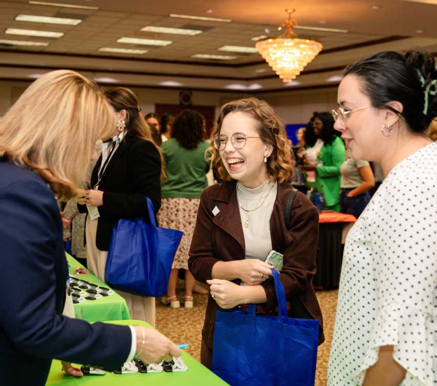 Two individuals with blue tote bags interact with a vendor at a table during an indoor event or conference.