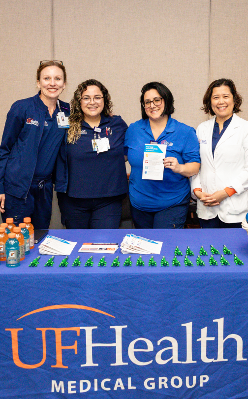Four individuals in medical attire behind UF Health Medical Group table with pamphlets, drinks, green items; indoor setting.