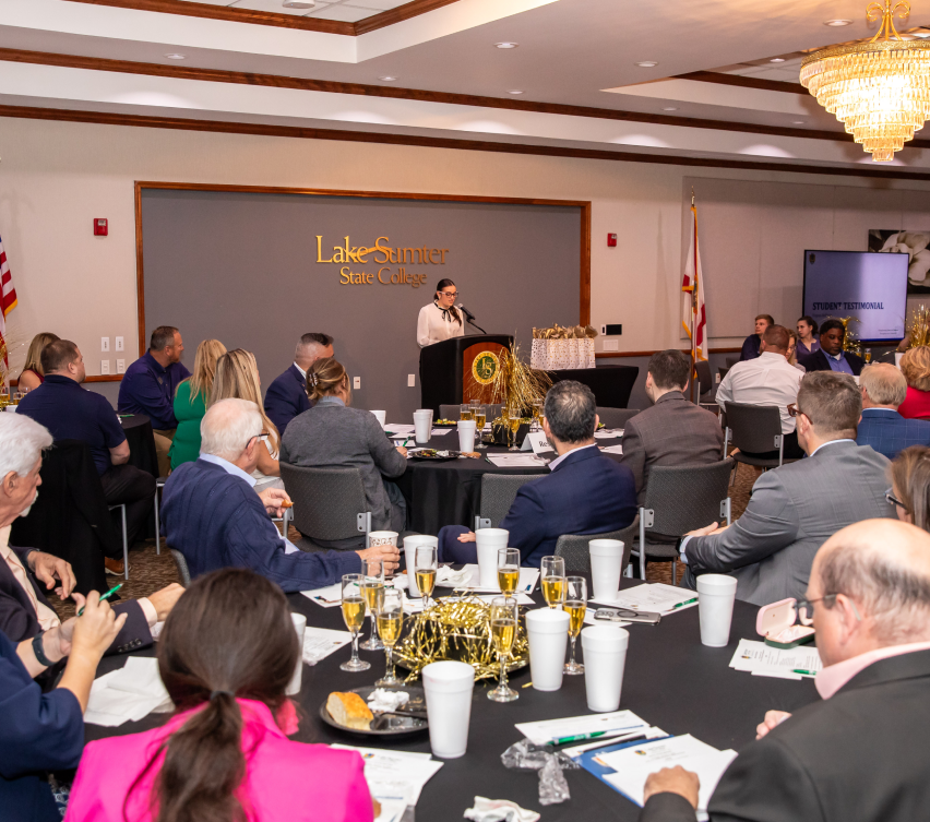 Individuals at round tables during formal event, person at podium speaking, Lake-Sumter State College backdrop, event setting.