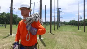 Person in orange long-sleeve shirt and hard hat carrying a coil of rope in a field of utility poles.