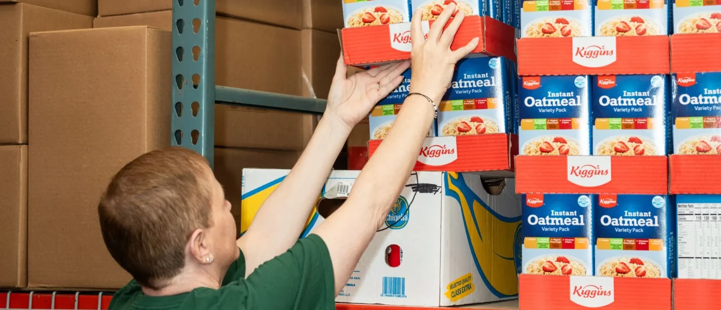 person reaches up to place or retrieve a box of oatmeal from a shelf in a warehouse. The shelves are stocked with similar oatmeal boxes, and large cardboard boxes are stacked nearby.