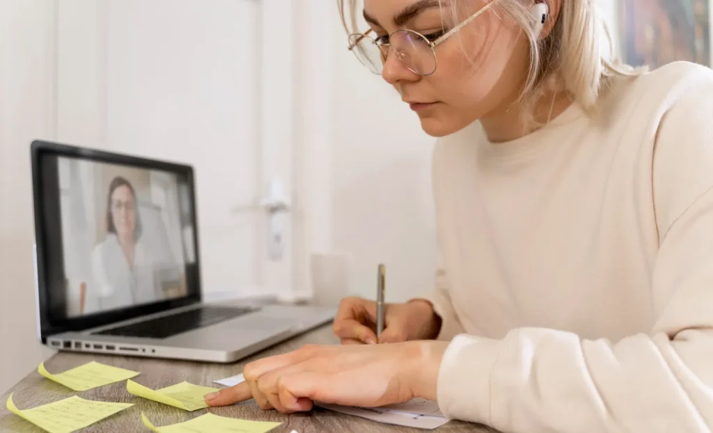 Individual taking notes during video call on laptop, with sticky notes on table; wearing glasses and earbuds. Setting unclear.