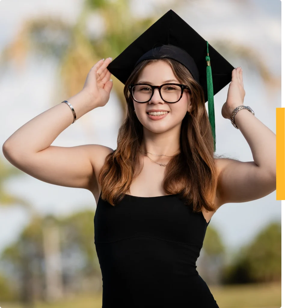 Student in black dress and graduation cap stands outdoors, smiling and holding cap with both hands.