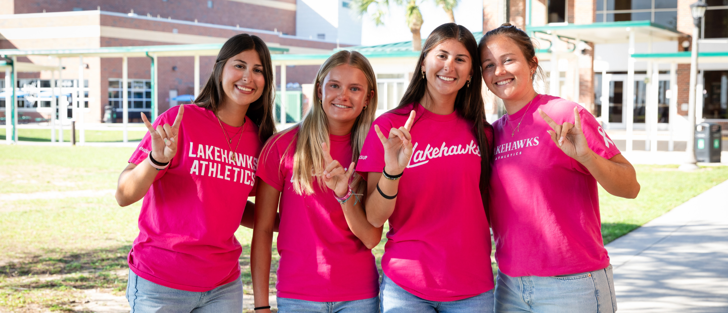 Four students in pink Lakehawks shirts stand outdoors, smiling and making hand signs.
