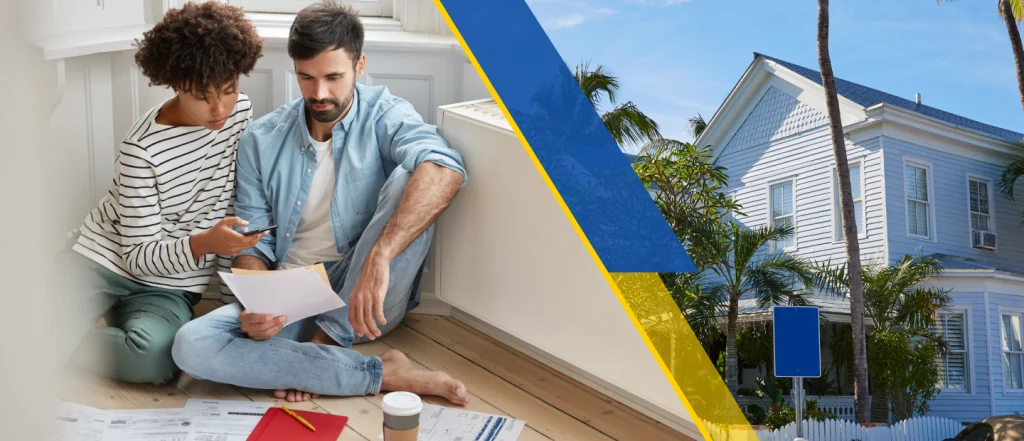 1 image files updated A man and woman sit on a floor looking at documents and a phone, with a white house and palm trees visible in the background
