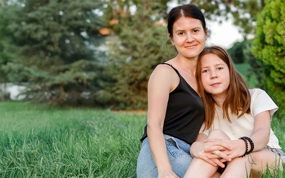 Alt text: Two people sit closely together on grass in a park-like setting. One wears a black sleeveless top and light blue jeans, while the other wears a white short-sleeved shirt and has their arms around the other's waist