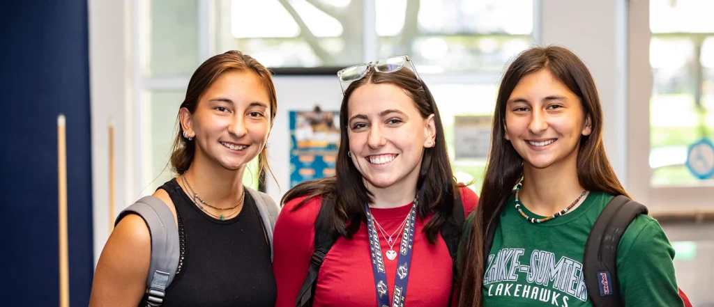 three female students standing together facing the camera. Their faces are blurred for privacy. The person on the left wears a black sleeveless top and a backpack. The middle person wears a red shirt, glasses on their head, and a lanyard. The person on the right wears a green 'Lake-Sumter Lakehawks' shirt and a backpack. Behind them are windows with visible posters