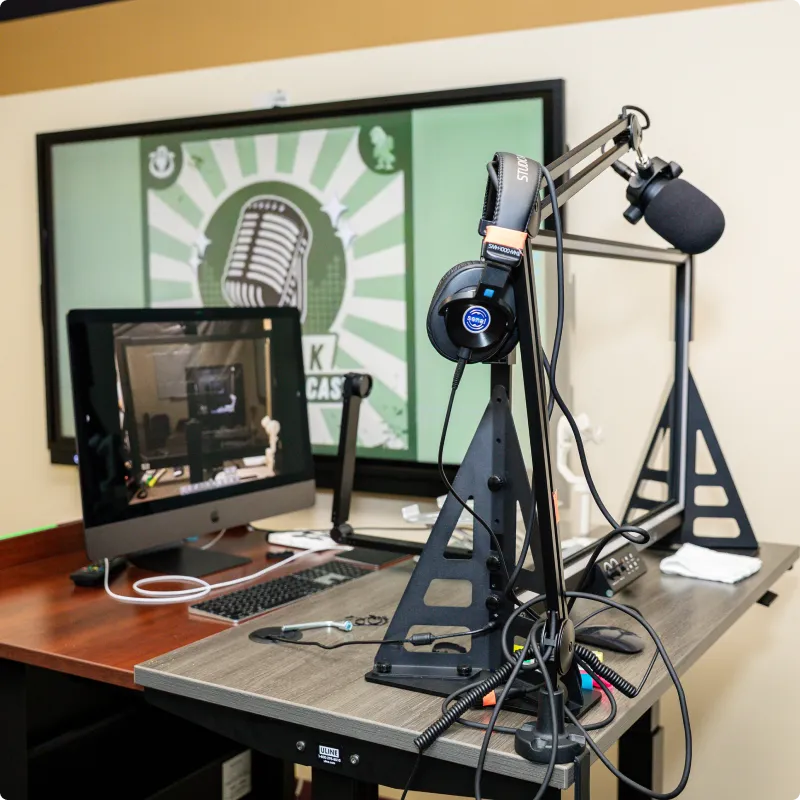 Podcast recording setup: microphone, headphones, computer on desk; podcast logo on wall in background.