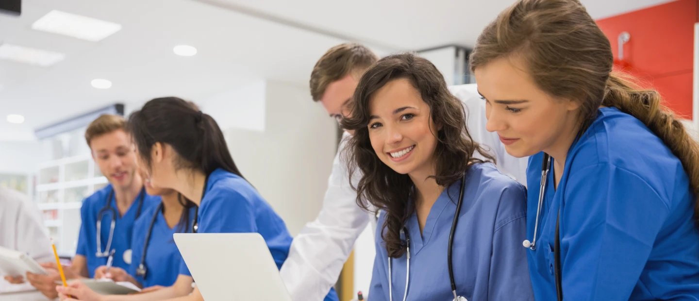 Smiling nurse in scrubs stands in a hospital hallway with other medical staff