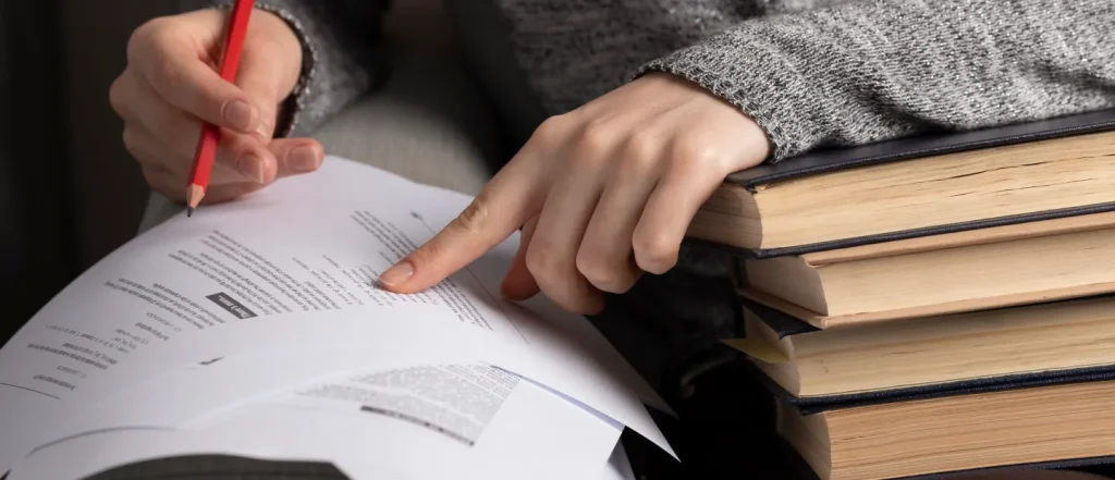 Person with red pencil reviewing documents, pointing at text, stack of books nearby. Likely individual studying.