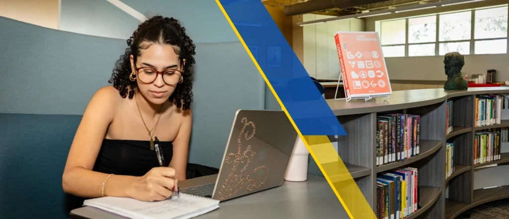A young woman with glasses and a black top is sitting at a table in a library, writing on a laptop