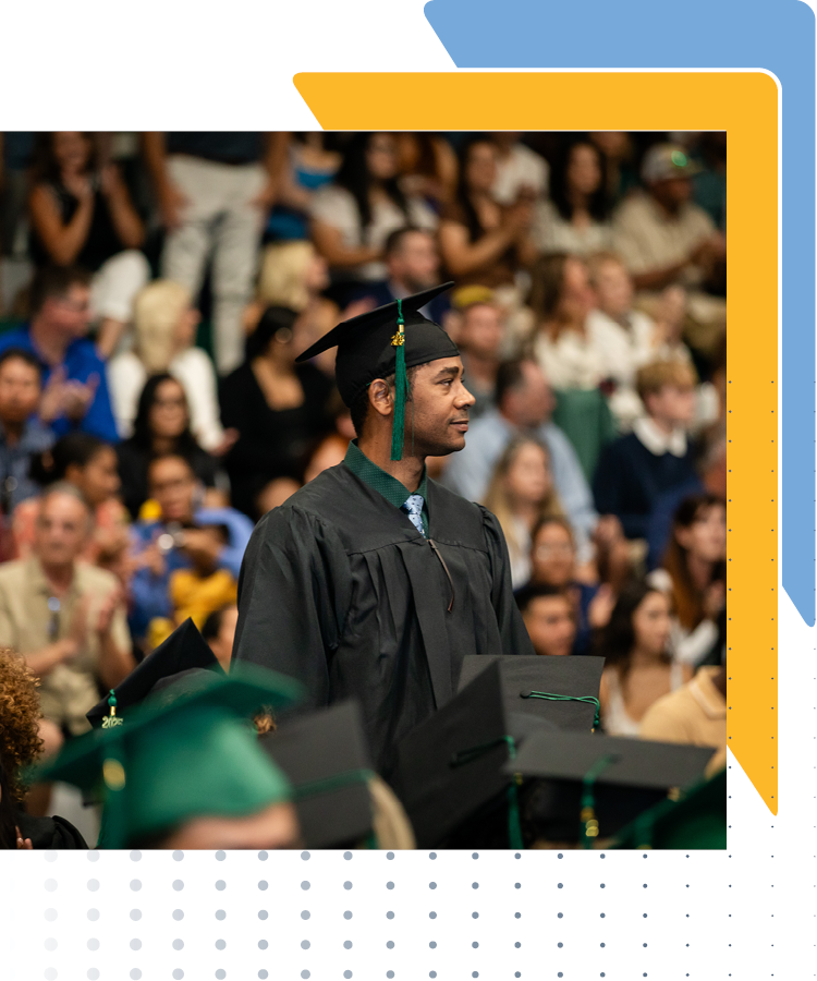 Grad student in a black gown and cap stands in a crowded auditorium.