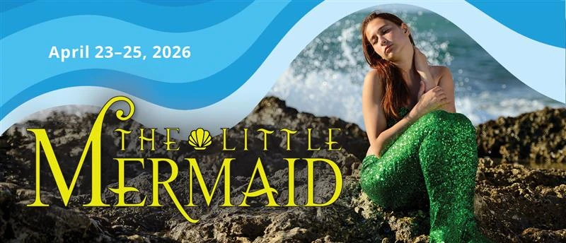 Student performer wearing a shimmering green mermaid costume with tail sits posed on dark rocks at the ocean's edge; waves splash against the rocks in the background.