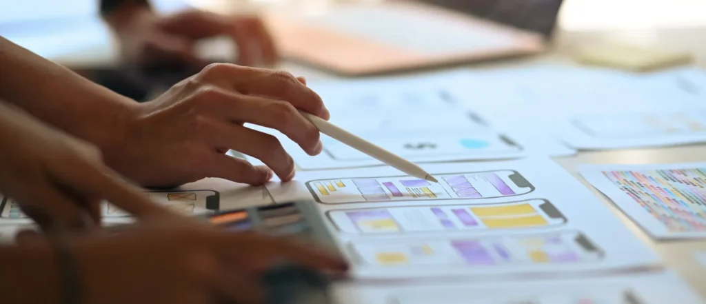 Hands point at smartphone app sketches on paper with color charts, emphasizing accessibility in design. Classroom or workspace setting.