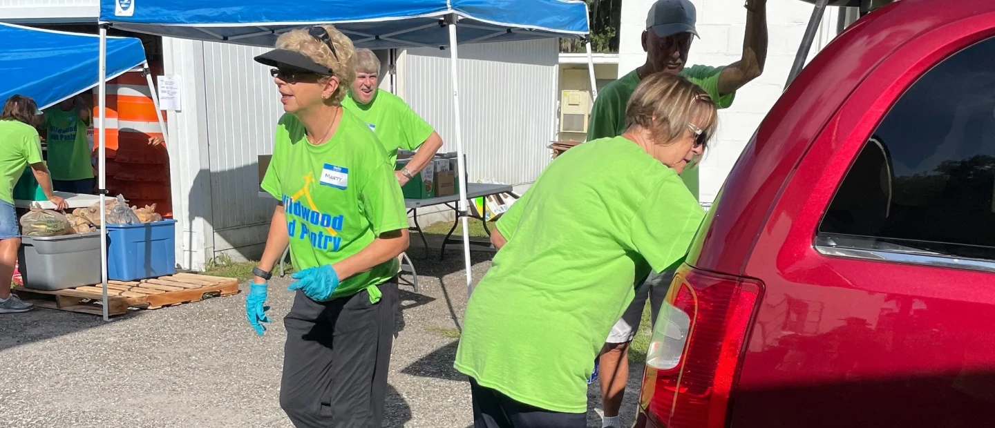 volunteers in green "Wildwood Food Pantry" shirts distribute food at outdoor tables near a red vehicle and blue tent